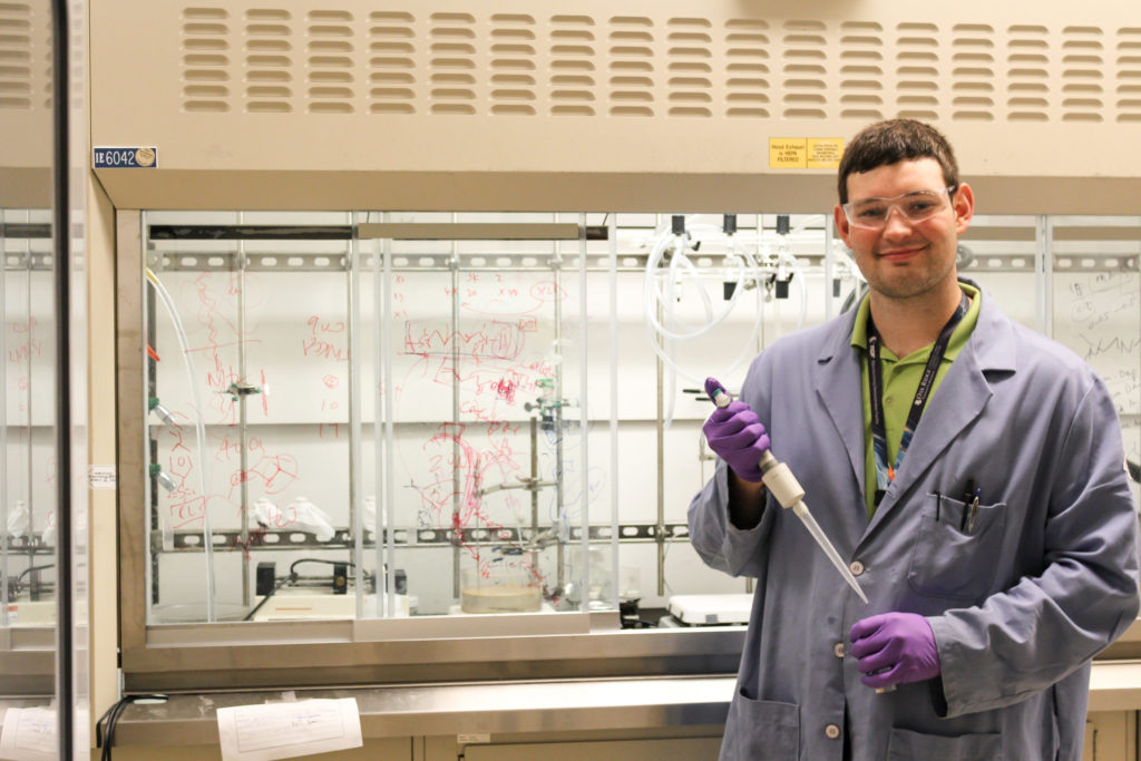 Man in purple lab coat holds instrument in a laboratory, smiling for the camera